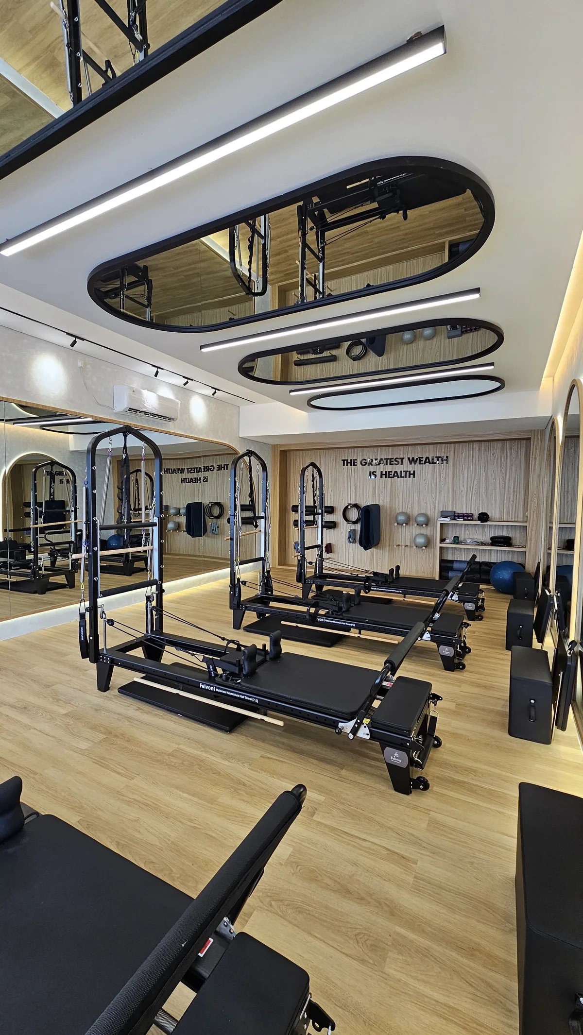 The studio: towers and reformers beneath oval ceiling mirrors, oak floor and a wall that reads The greatest wealth is health